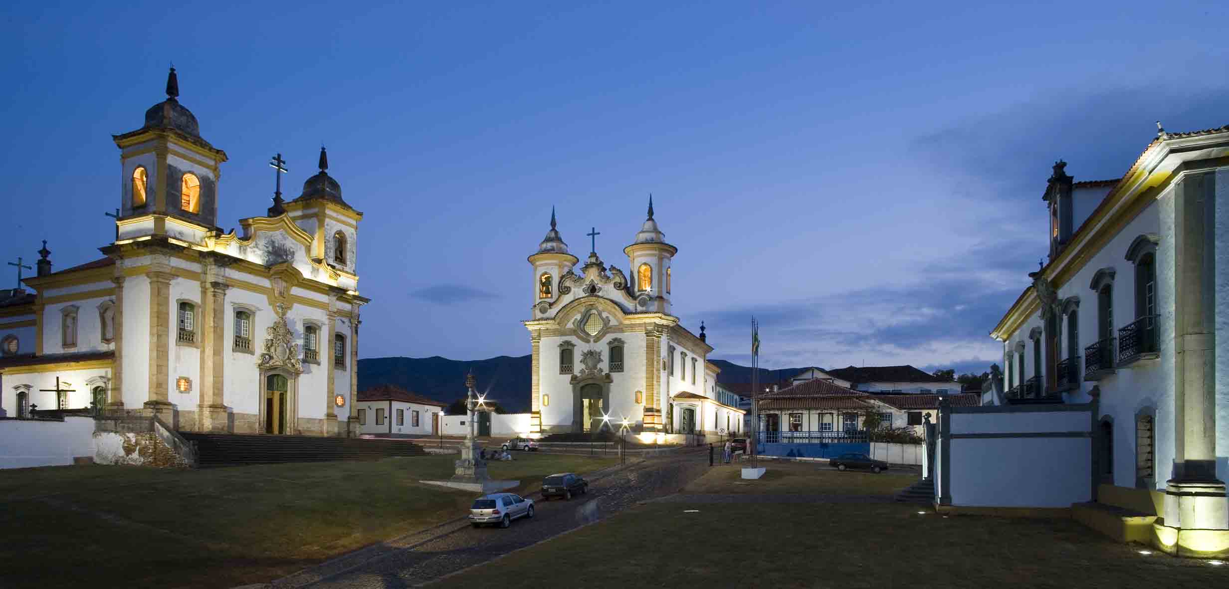 Igrejas de São Francisco de Assis e do Carmo iluminadas ao anoitecer, em praça de Mariana